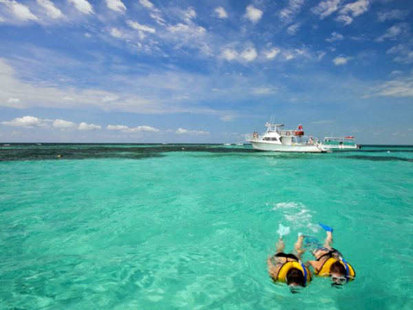 Two people snorkeling in clear turquoise water with a boat in the distance under a partly cloudy blue sky.