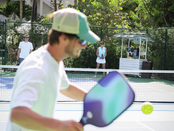 A person is playing pickleball on an outdoor court, holding a paddle, with a ball in motion, while others watch in the background.