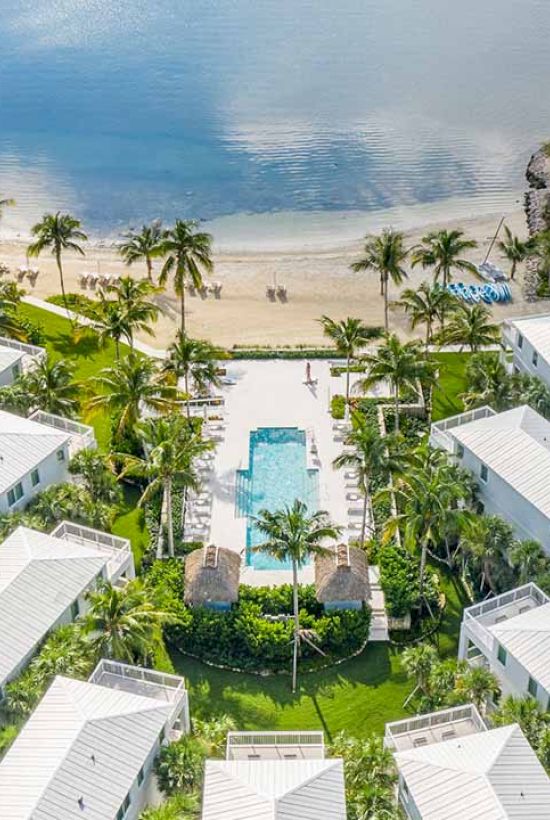 Aerial view of a beachside resort with white buildings, a central pool, lush greenery, and a sandy beach leading to calm waters in the background.