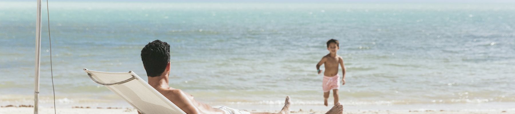 A person relaxes on a lounger under an umbrella at the beach, while a child approaches. The sky and sea are calm and picturesque.