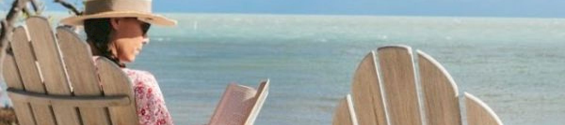 A person in a hat is sitting on a beach in a chair, reading a book, with the sea and sky in the background.