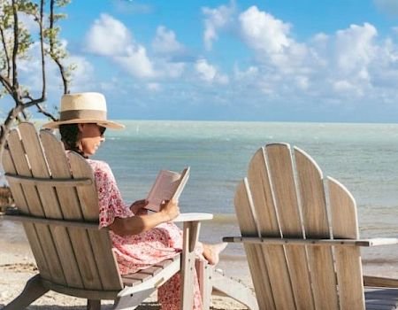A person in a hat is sitting on a beach in a chair, reading a book, with the sea and sky in the background.