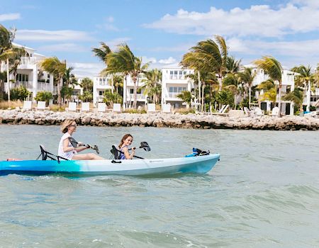 Two people are kayaking in a blue and white kayak on the water, with modern white buildings and palm trees visible in the background.