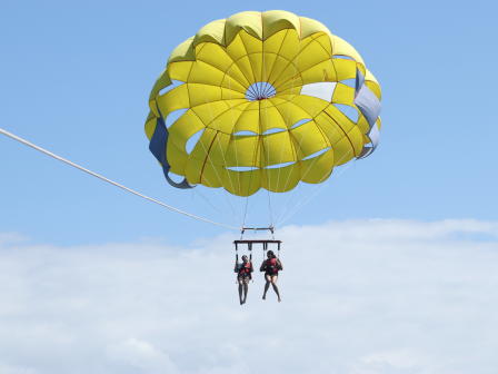 Two people are parasailing with a bright yellow parachute against a clear blue sky.
