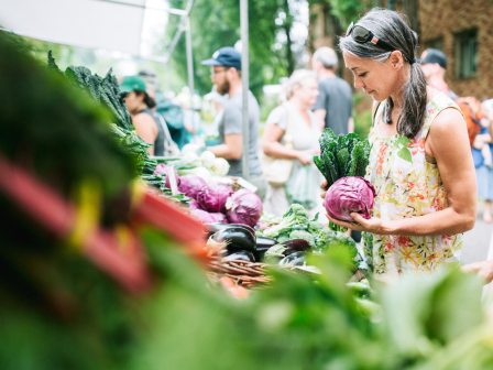 A woman is selecting red cabbage at a farmers' market stall, surrounded by various fresh vegetables and shoppers.
