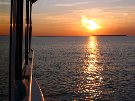 A serene sunset over the ocean, viewed from the side of a boat, with golden reflections on the water's surface.