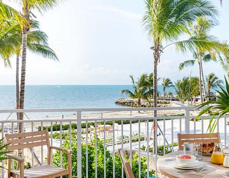 A tropical beachfront view with palm trees, a patio table set for breakfast, and a railing overlooking the ocean on a sunny day.