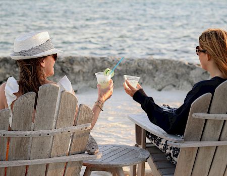 Two people sitting on wooden chairs by the beach, holding drinks and facing the ocean, enjoying a relaxing moment.