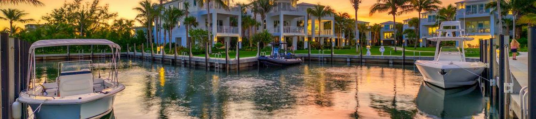 A peaceful marina scene with boats docked, surrounded by palm trees and buildings, under a stunning sunset sky reflected in the water.