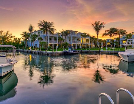 A peaceful marina scene with boats docked, surrounded by palm trees and buildings, under a stunning sunset sky reflected in the water.