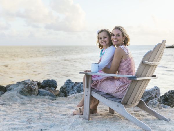 Two women sit close together on a beach chair by the water, smiling as they enjoy a sunny, relaxing seaside moment.