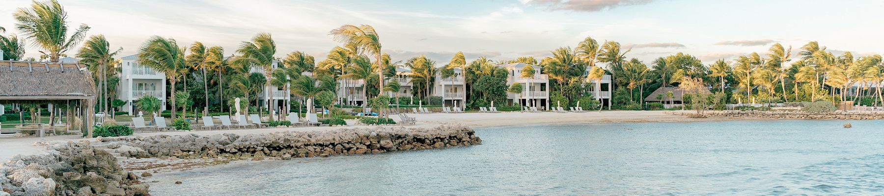 A scenic coastal view with palm trees, houses, and a calm sea under a blue sky with clouds, along a rocky shoreline.