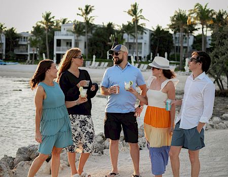 A group of people stand on a beach, chatting and holding drinks, with palm trees and buildings in the background.