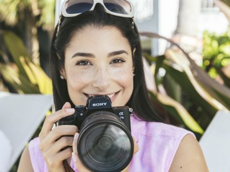 A person wearing sunglasses on their head smiles while holding a Sony camera outdoors, with plants and a building in the background.
