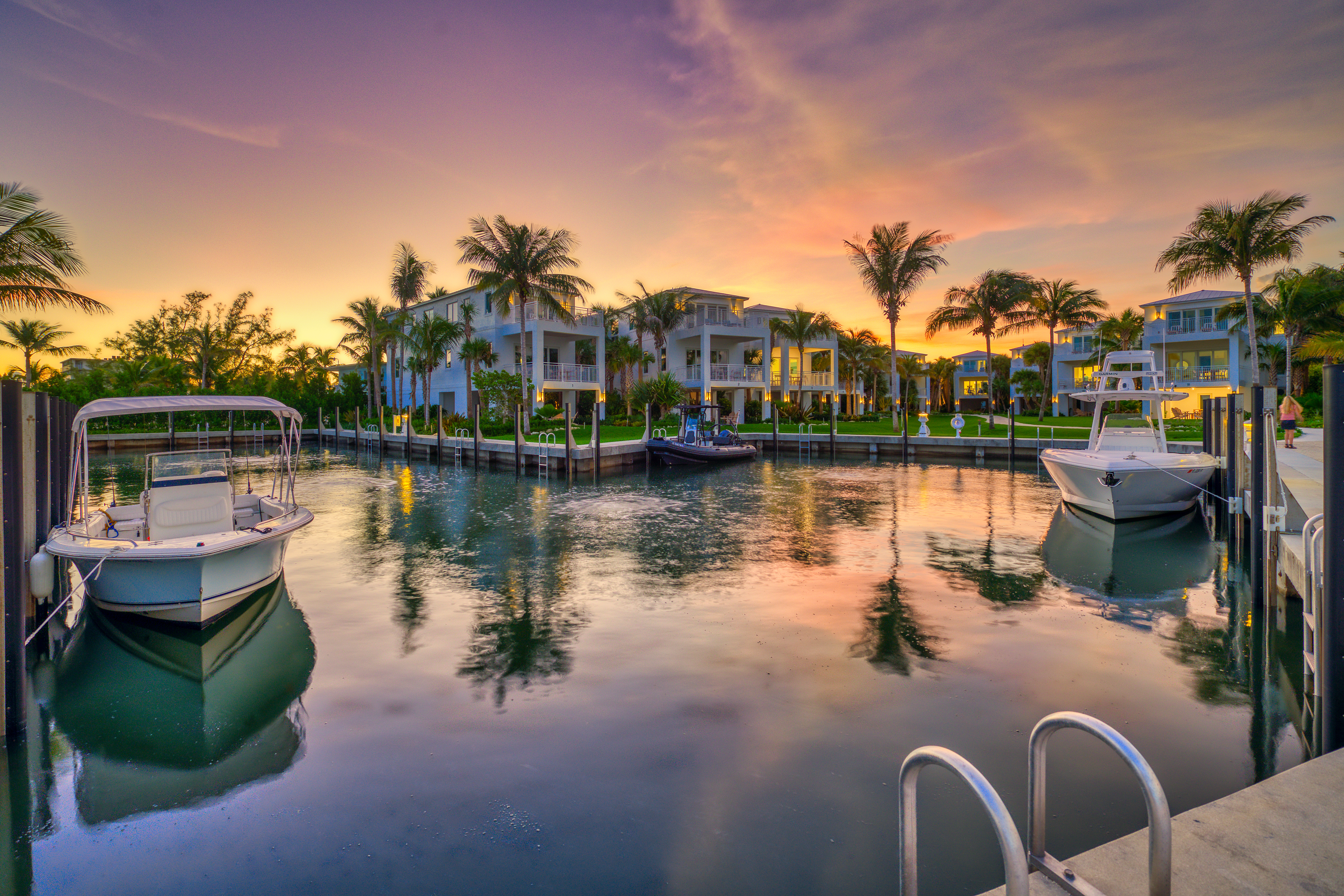 A serene marina at sunset with palm trees, docked boats, and luxury houses reflected in the calm water.