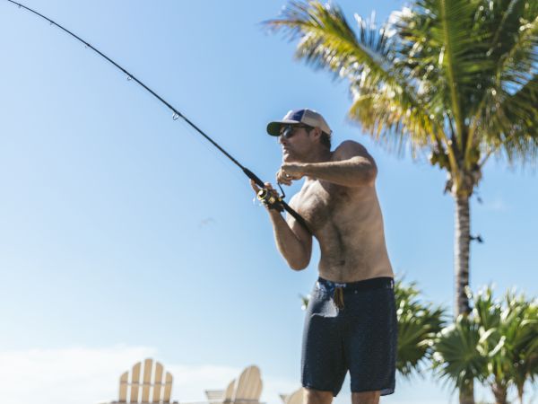 A person is fishing on rocky terrain with a palm tree and Adirondack chairs in the background under a clear blue sky.