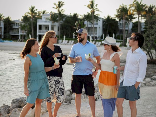 A group of people stand on a beach, chatting and holding drinks. Palm trees and houses can be seen in the background.