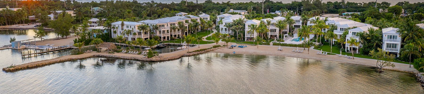 A coastal view at sunset with buildings, palm trees, and calm water under a colorful sky.
