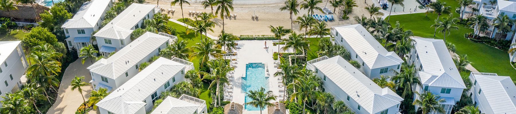 Aerial view of a beachfront resort with white buildings, a central pool, and surrounding palm trees, adjacent to a serene body of water.