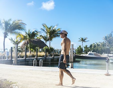 A person wearing a cap and shorts walks along a sunny dock lined with palm trees and boats, under a clear blue sky.