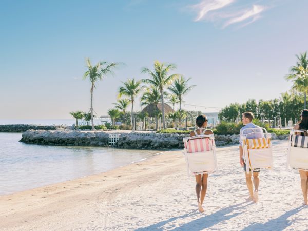 Three people walk along a sandy beach carrying striped bags, with palm trees and calm water in the background.
