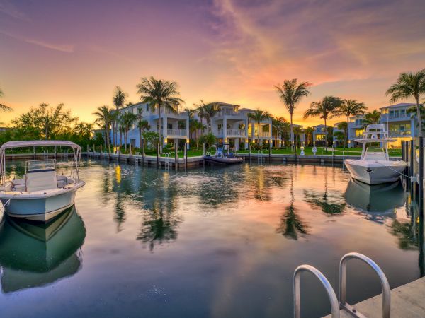 Sunset over a calm marina with boats, palm trees, and reflections on the water, as houses line the shore.