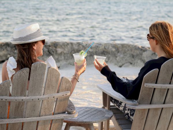 Two people sit in wooden chairs by the beach, holding drinks, enjoying the view and relaxation together.