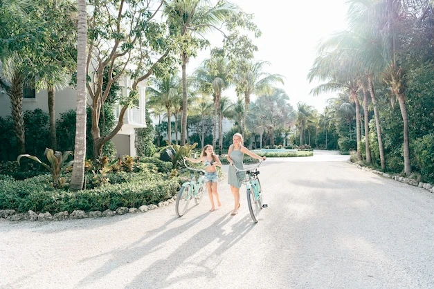 Two people stand with bicycles on a sunny, palm-lined path near a gardened condo entrance, smiling as they pose for a photo.
