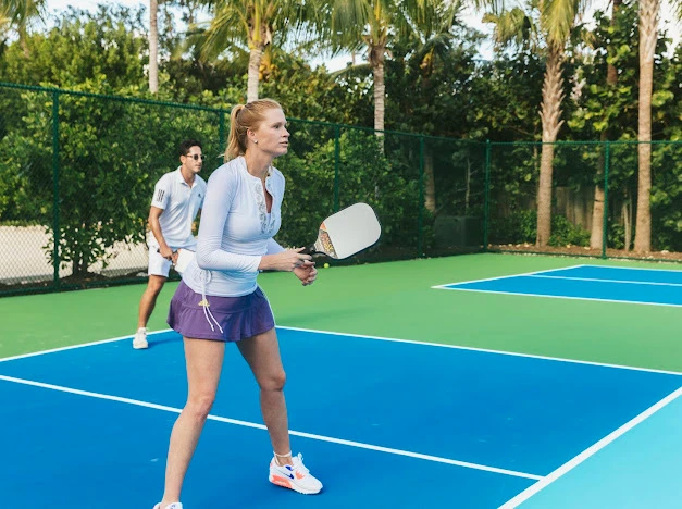 Two people play pickleball on a blue court; a woman in a light blue top and purple shorts holds a paddle, ready to return.
