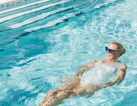 A person floating on their back in a clear blue pool, wearing sunglasses, sunlit water shimmering around them.