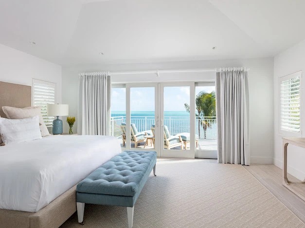 A bright hotel room with a king bed, tufted blue bench, soft neutrals, and sliding glass doors opening to a balcony with ocean view.