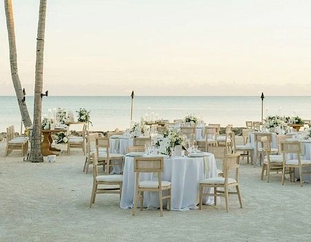 Beach wedding setup on white sand with palm trees, string lights, and many round tables with white linens facing the ocean.