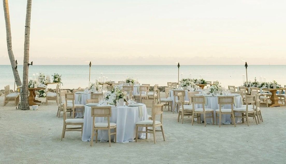 Beach wedding setup on white sand with palm trees, string lights, and many round tables with white linens facing the ocean.