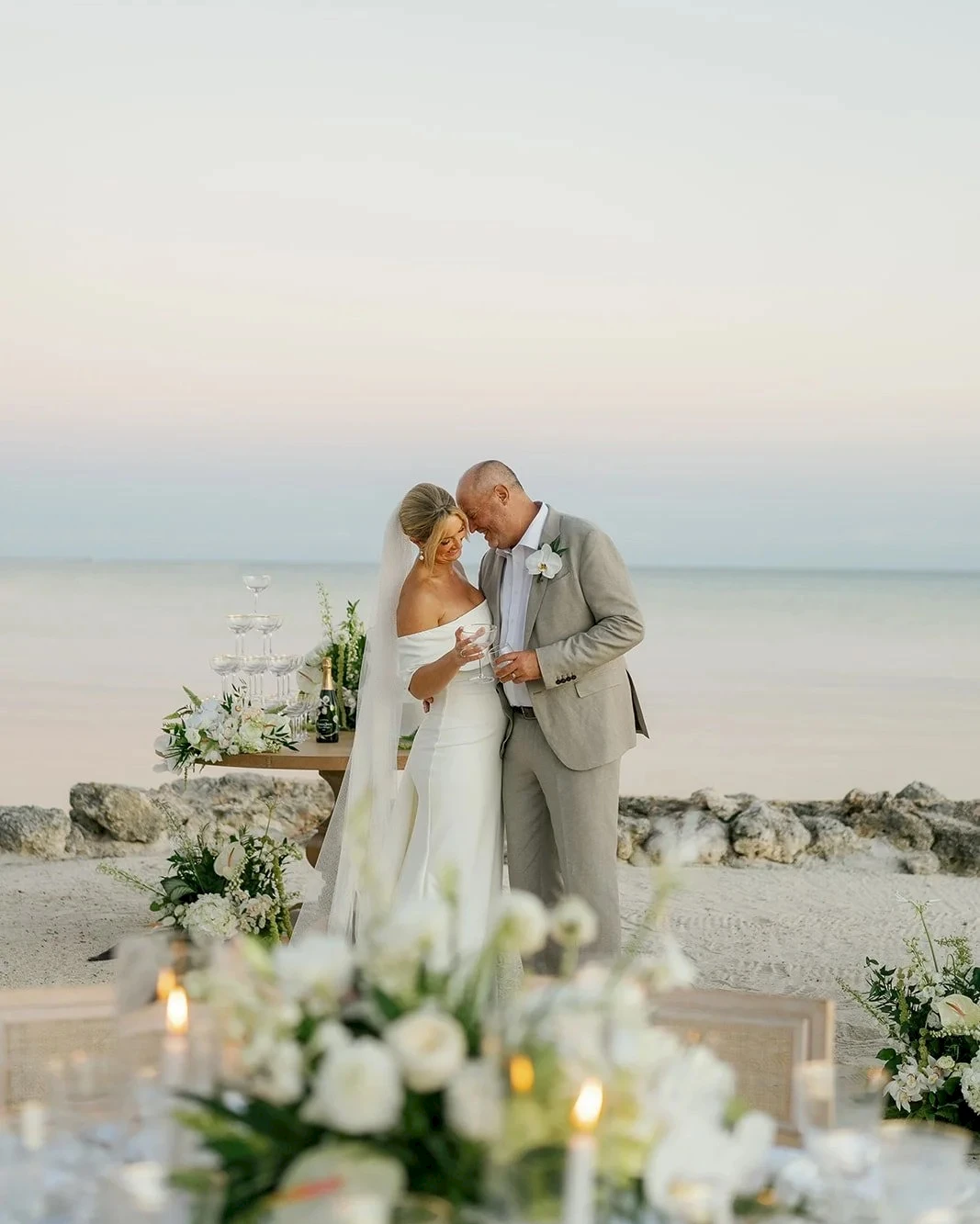 A wedding couple kisses by the sea at sunset, with a floral table setup and soft, romantic decor along a rocky shoreline. end.