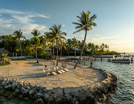 Palm-fringed tropical beach with lounge chairs, a rocky jetty, and calm blue water at sunset; a tranquil island paradise.
