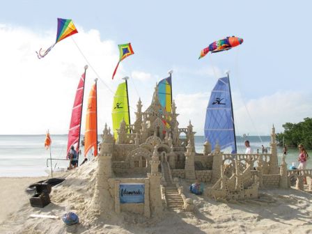 A large sandcastle on the beach with colorful kites and sails, people playing nearby, and the ocean in the background.