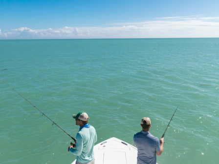 Two people are fishing from a boat on calm turquoise coastal waters under a clear blue sky.