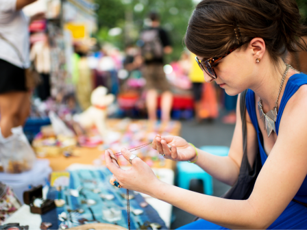 A person in sunglasses examines jewelry at a colorful outdoor market, surrounded by various items on display on a table.