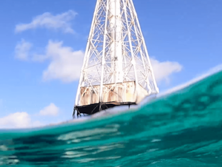 A lighthouse tower rises above the waterline as waves splash around, with a diver or swimmer visible underwater near the bottom, in clear blue seas.