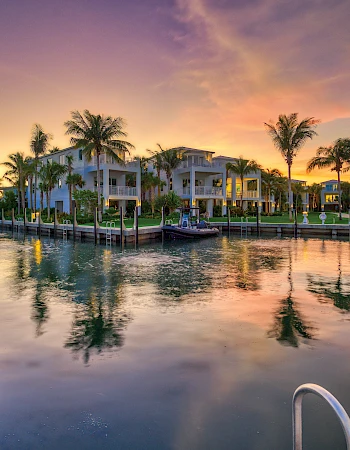 Sunset over a quiet marina with boats, palm trees, and pastel houses along calm water, reflections shimmering on the rippling surface.