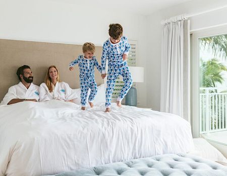 Two kids jump on a bed with parents nearby in white robes; bright room, balcony door, sunny and cheerful vibe.