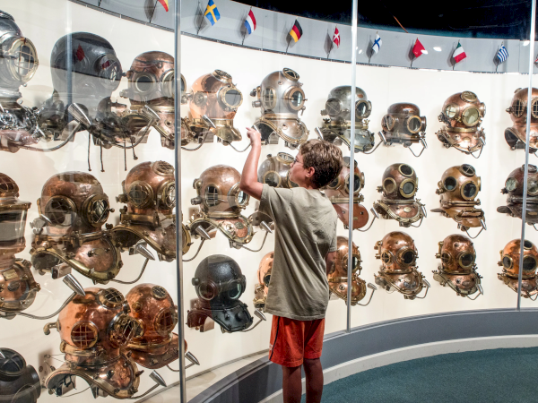 A boy in a museum reaches for vintage military helmets displayed on glass shelves, with many helmets of various colors and shapes, flags above.
