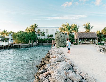 A rocky jetty extends into calm turquoise water, with palm trees, white buildings, and a thatched shelter in a sunny coastal scene.