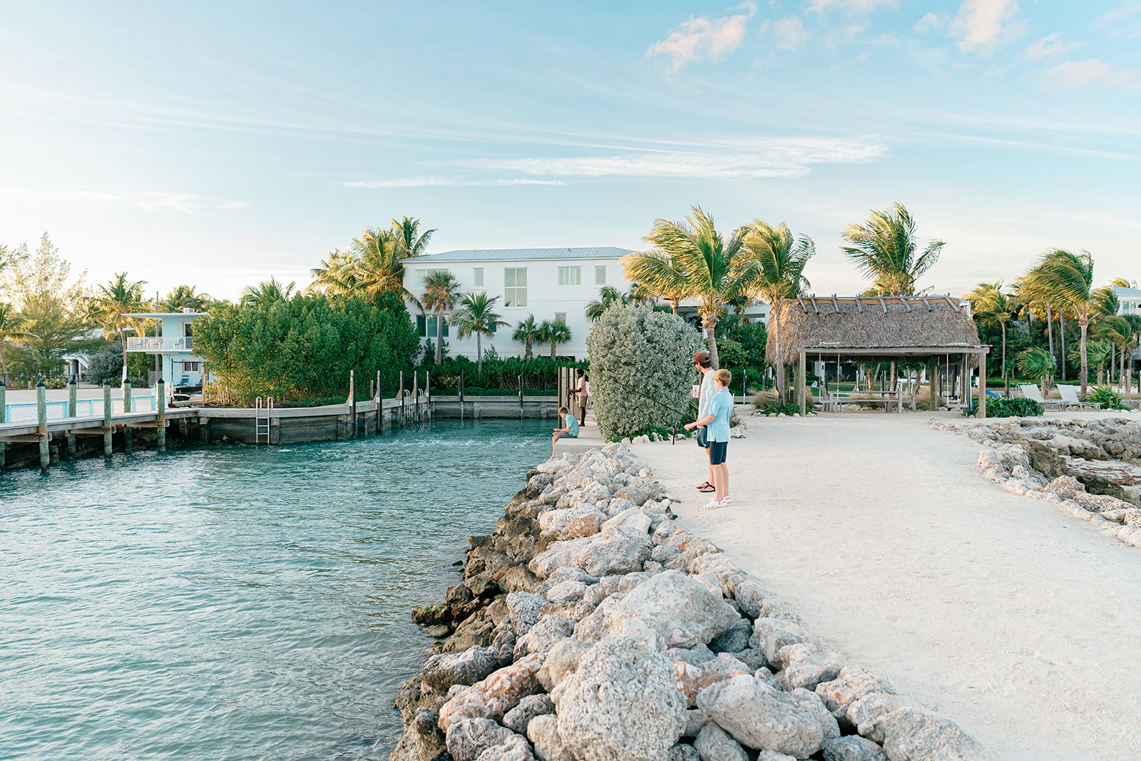 A rocky jetty extends into calm turquoise water, with palm trees, white buildings, and a thatched shelter in a sunny coastal scene.