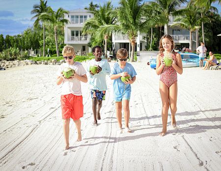 A group of kids walking on a sunny sandy beach, holding coconuts. Palm trees and a resort in the background; everyone looks happy.