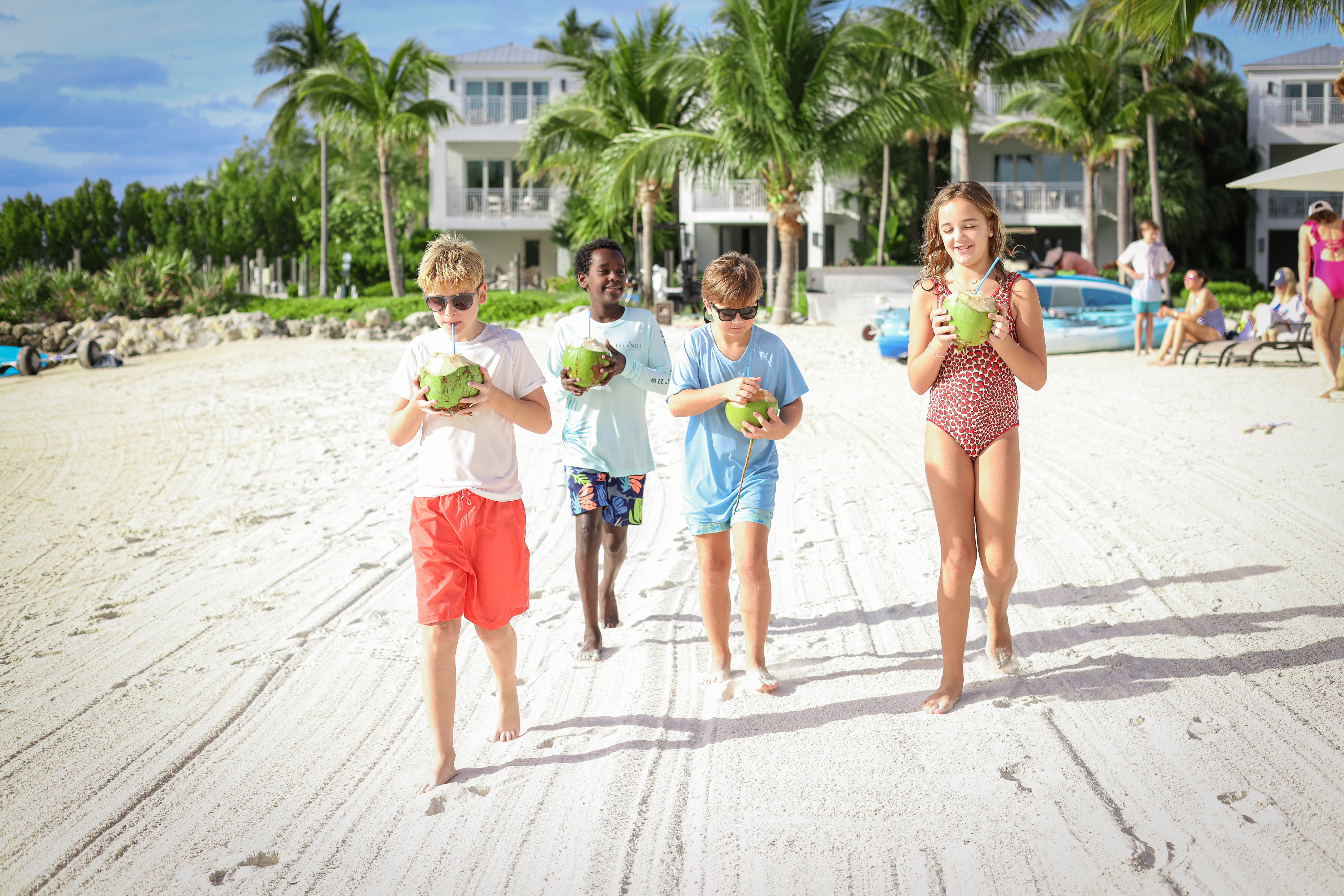 A group of kids walking on a sunny sandy beach, holding coconuts. Palm trees and a resort in the background; everyone looks happy.