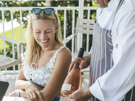 A smiling woman in a floral top sits on a balcony as a waiter pours ros&eacute; from a bottle labeled "ROS&Eacute;" beside her.