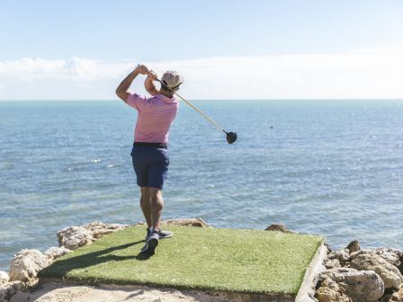 A golfer tees off on a rocky cliff, swinging over the sea, with a clear blue sky above, on a small grassy mat by the water.