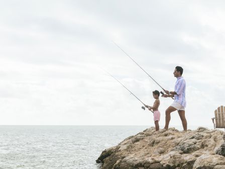 Two people, a man and a child, fishing off a rocky coastline at the edge of the water, with a chair nearby and a calm sea under a cloudy sky.