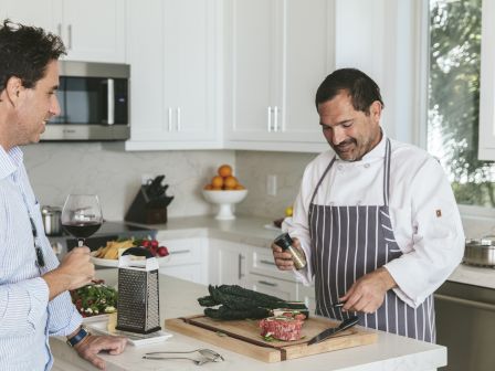 Two men in a bright kitchen: one chef in a striped apron seasons meat on a cutting board while a guest drinks red wine and chats, all to relax.